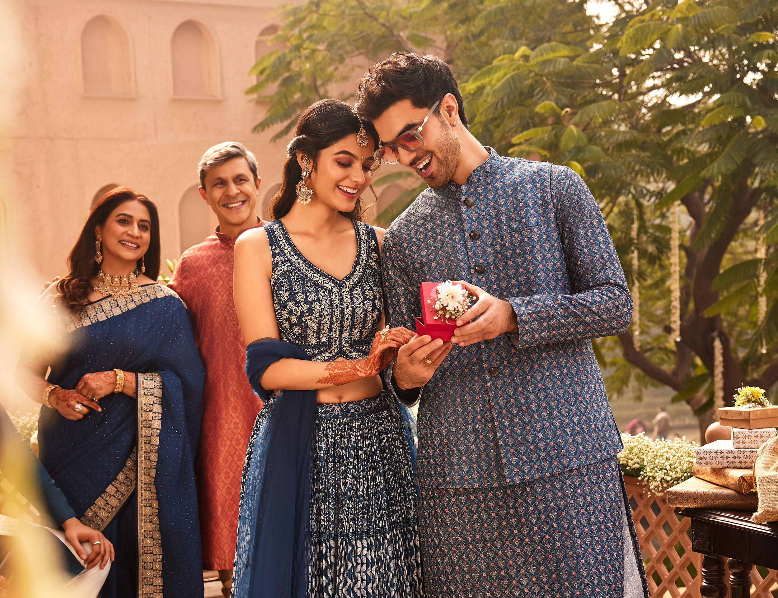 Smiling couple in coordinated blue traditional Indian attire exchanging a gift, with family members in festive ethnic wear standing beside them in an outdoor celebration setting.